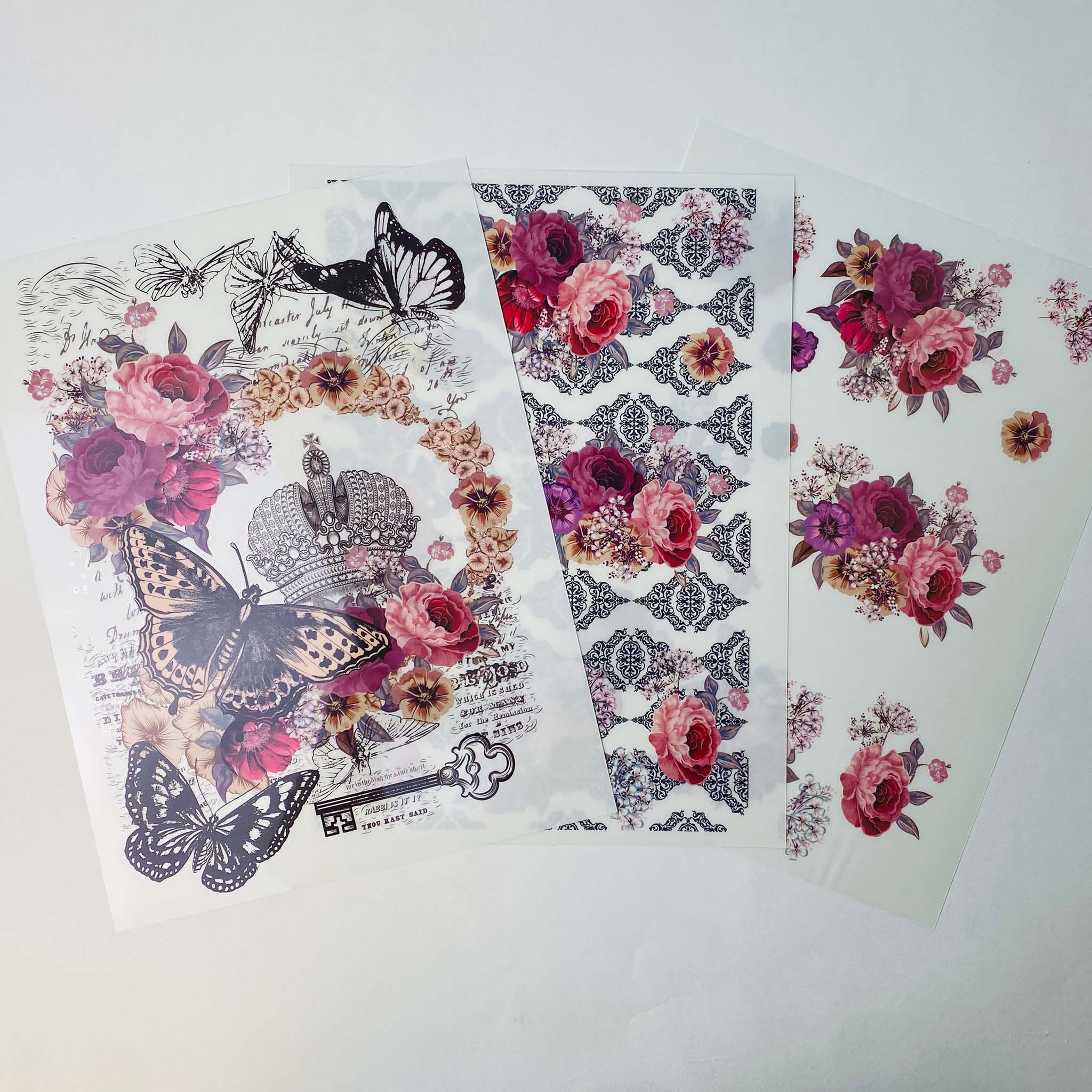 Three sheets of rub-on transfers that feature an ornate crown, romantic roses and fluttering butterflies; small bouquets of mauve and purple flowers; and a repeating black damask pattern with small bouquets of mauve and purple flowers.