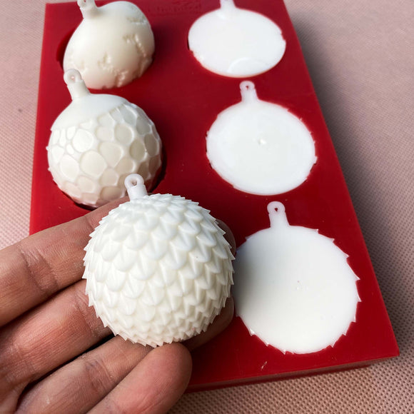 A red silicone mold and white resin castings of 6 half ornament bulbs are against a beige background. A hand is shown holding one of the castings.