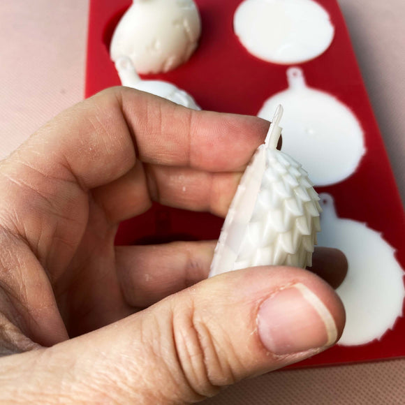 A red silicone mold and white resin castings of 6 half ornament bulbs are against a beige background. A hand is shown holding one of the castings.