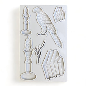 A light grey silicone mold of stacks of books, candles, a branch, and a bird is against a white background.