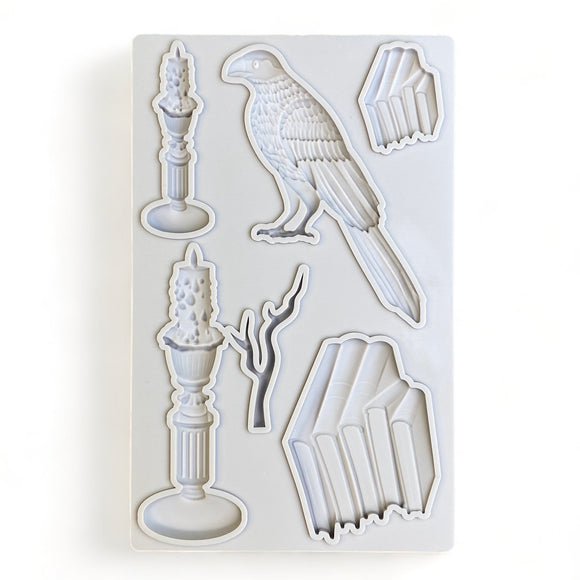 A light grey silicone mold of stacks of books, candles, a branch, and a bird is against a white background.