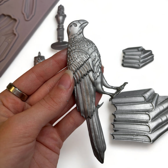 A light grey silicone mold and silver colored castings of stacks of books, candles, a branch, and a bird are against a white background. A hand is shown holding the bird.