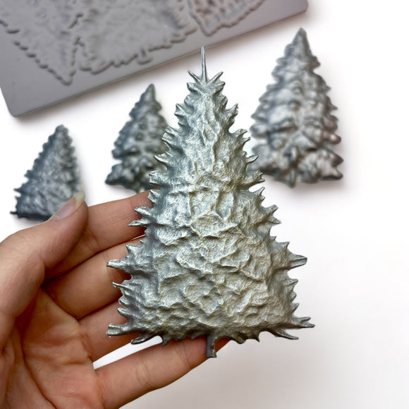 A beige silicone mold and silver colored castings of 5 varying size pine Christmas trees are against a white background. A hand is shown holding one of the castings.