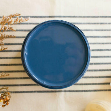 An arial view of an open container of Dixie Belle Paint Company's Cape Current All-In-One Silk Paint is on a white knit material with dark grey stripes.