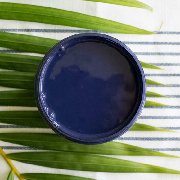 An arial view of an open container of Dixie Belle Paint Company's Nautical All-In-One Silk Paint is on a white material with dark grey stripes and a palm frond.