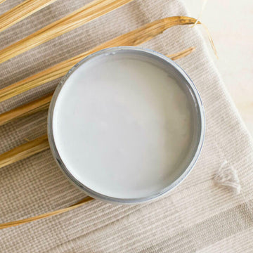 An arial view of an open container of Dixie Belle Paint Company's Quiet Cove All-In-One Silk Paint is on a white material with dried palm frond.