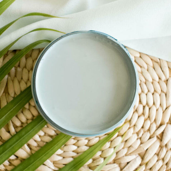 An arial view of an open container of Dixie Belle Paint Company's Nautical All-In-One Silk Paint is on a weaved wicker placemat with a white cloth napkin and green palm frond.