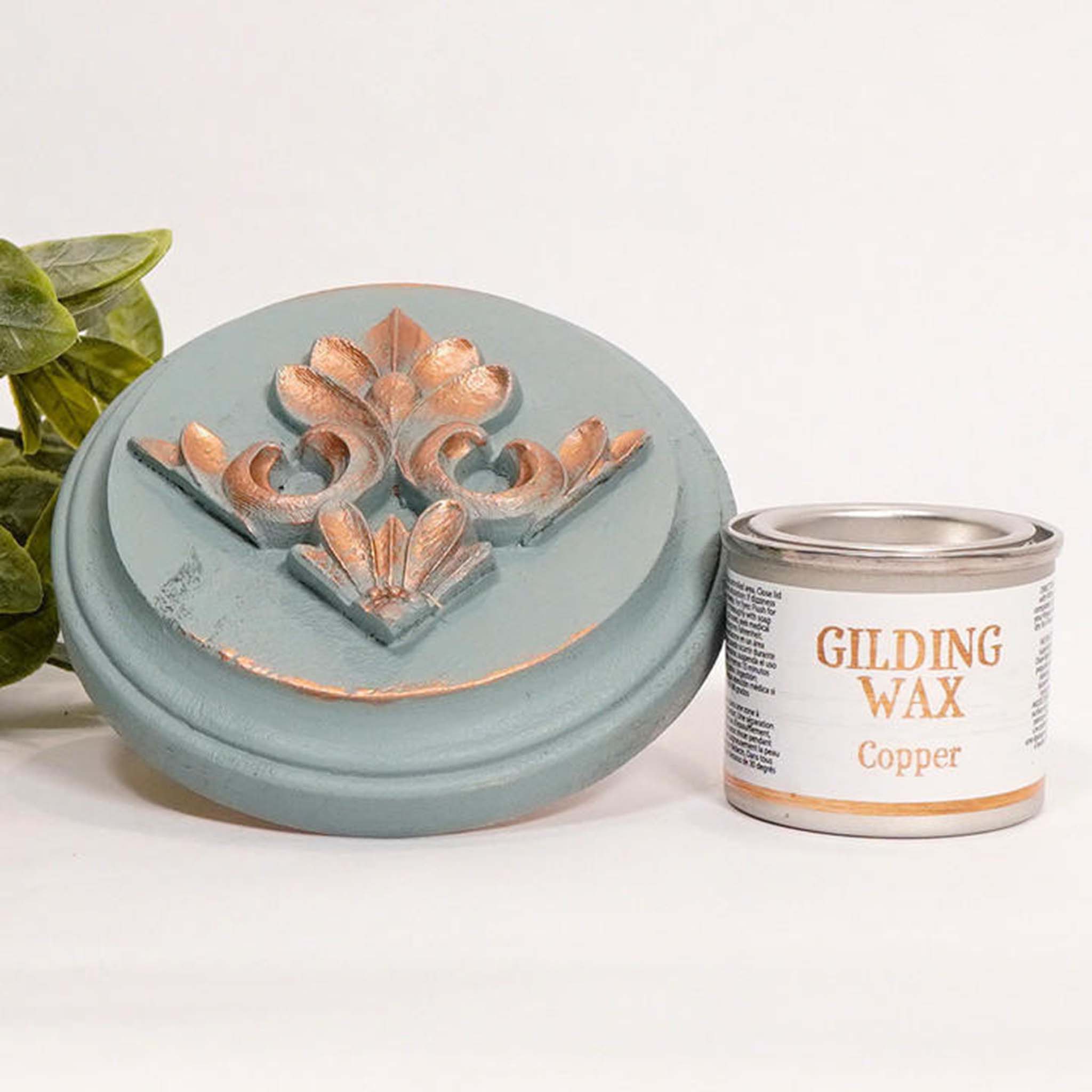 A can of Dixie Belle's Copper Gilding wax is next to a light blue painted round wood sample. The wood sample has an ornate silicone mold casting that features the copper gilding wax on it.