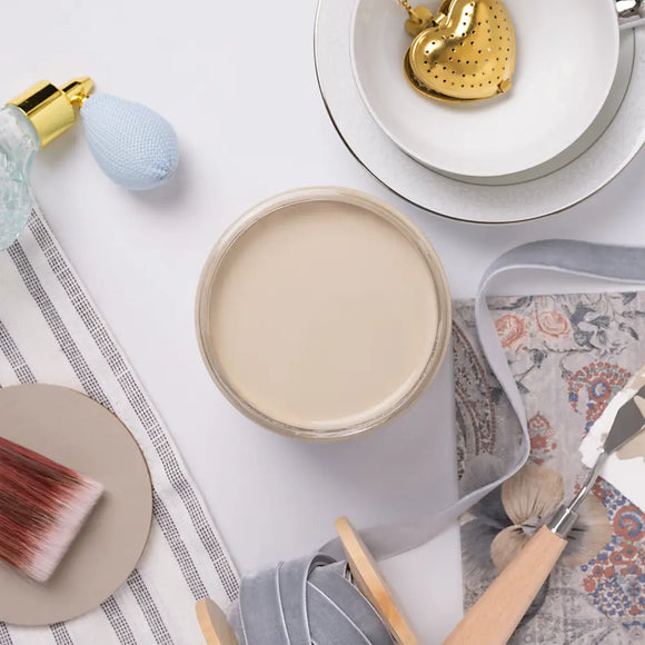 Jar of cream paint on a white surface with a brush and bowl in the background
