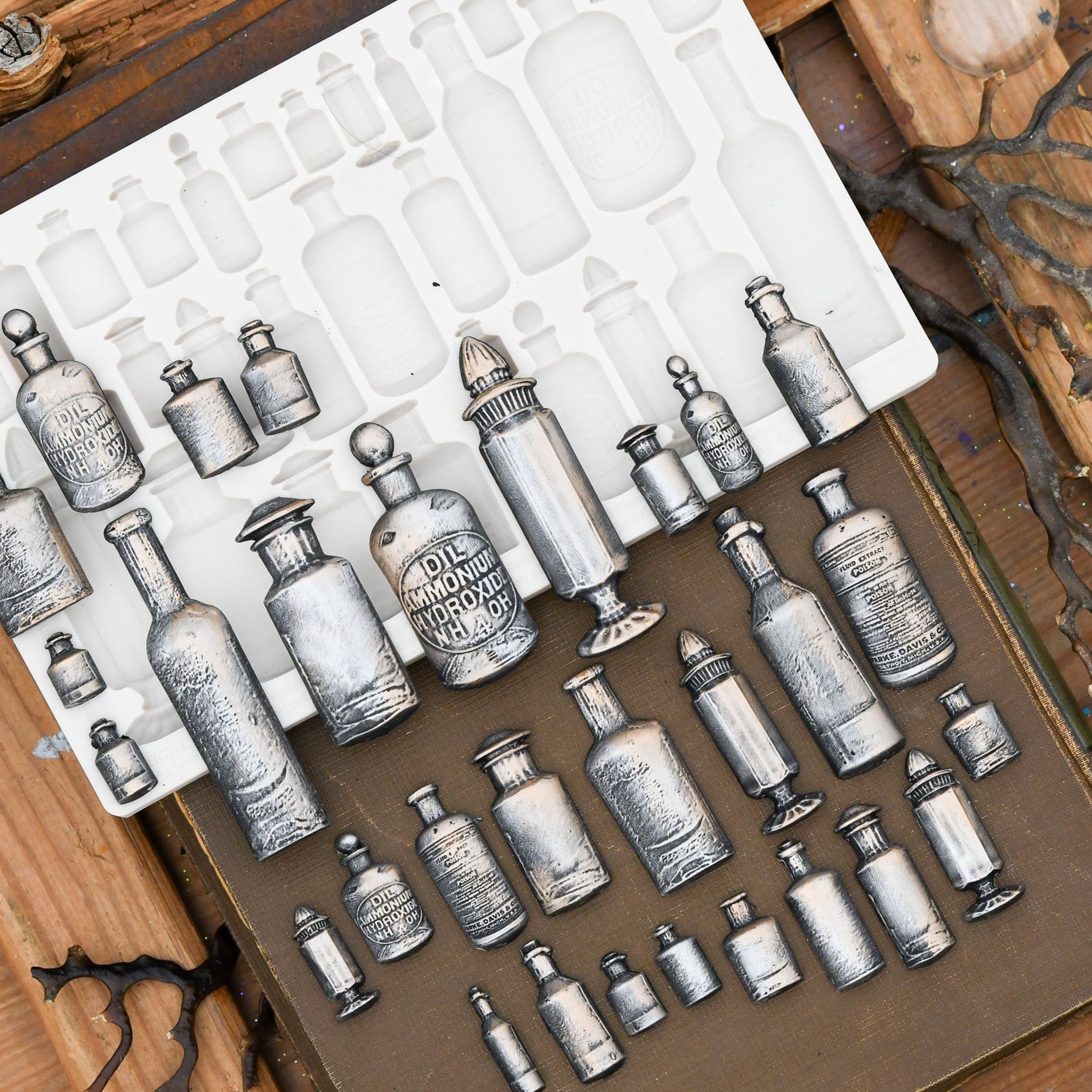 A beige silicone mold and silver-colored castings of 30 varying sizes of apothecary bottles are on top of a brown book against a wood background.