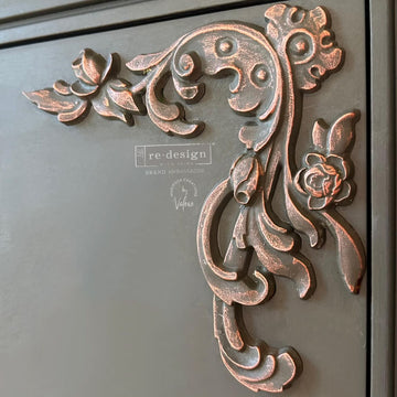 A close-up of a bronze wax accented ReDesign with Prima Intricate Grace Decor Poly Casting is shown on a black piece of furniture.