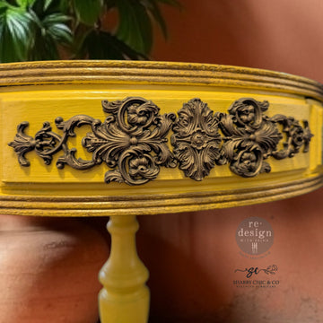 A close-up of a round vintage side table refurbished by Shabby Chic & Company is painted yellow and features ReDesign with Prima's Luxe Ornamenta Decor Poly Casting on the side.