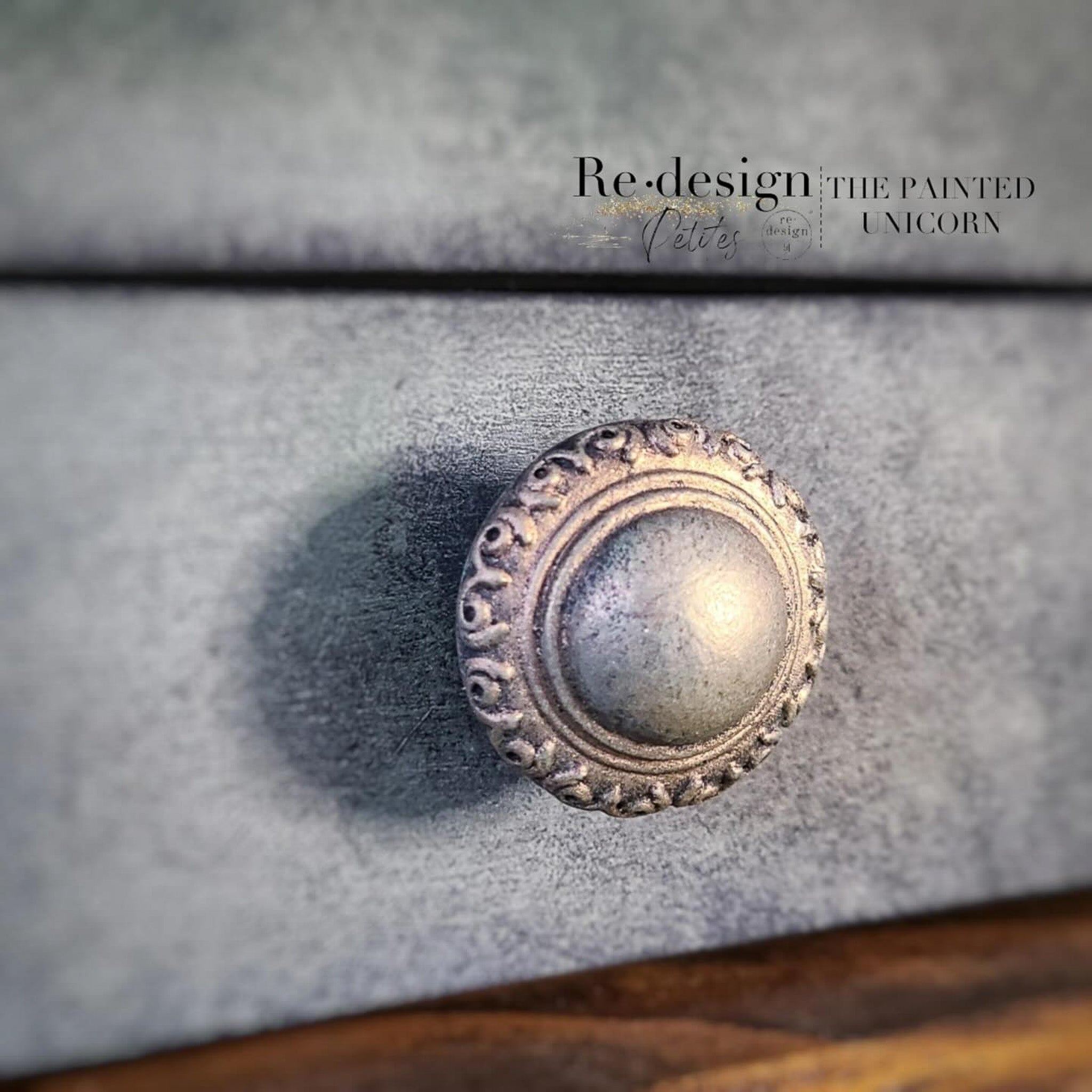 A close-up view of a round drawer knob with an ornate edging design created by The Painted Unicorn using Redesign with Prima's Luxe Ornate 3D Knob silicone mold.