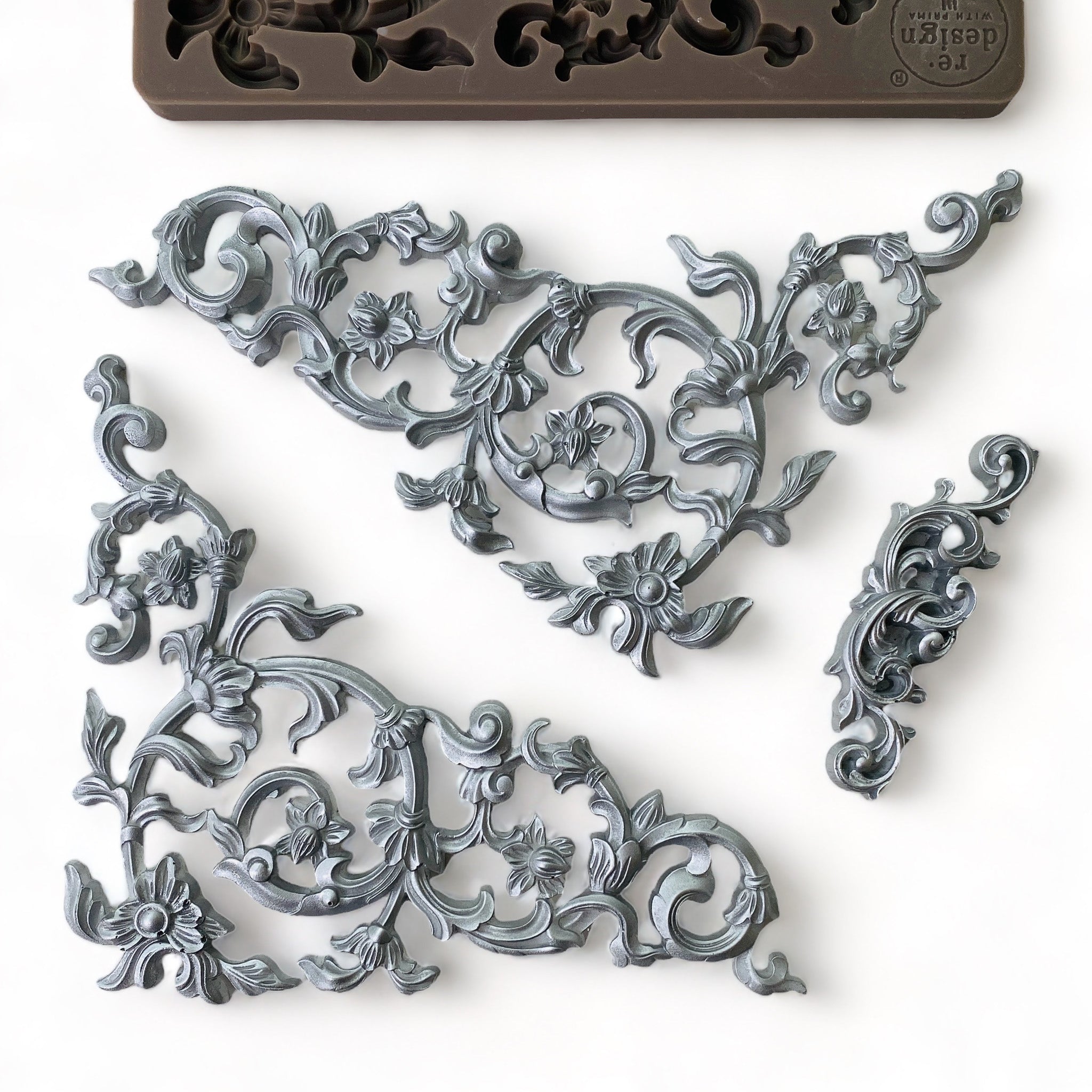 A brown silicone mold and silver-colored castings that feature 2 scrolling vine corners and a small center accent scroll are against a white background.