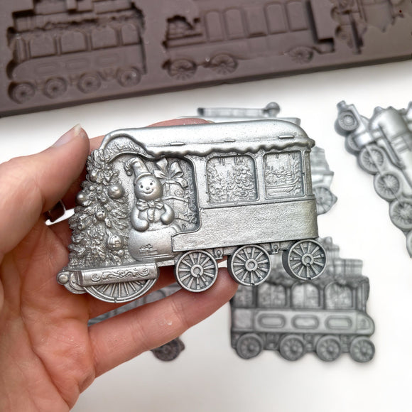 A close-up of a hand holding a silver colored casting made from ReDesign with Prima's The Jolly Express silicone mold is against a white background.