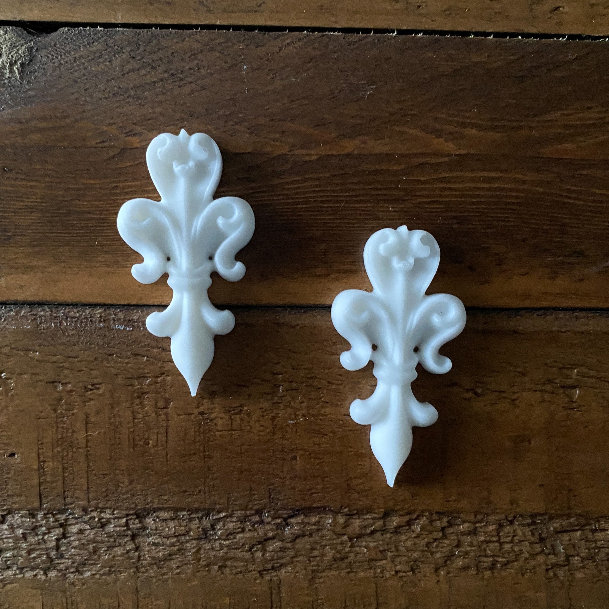 Two small white resin casted fleur de lis are against a wood background.