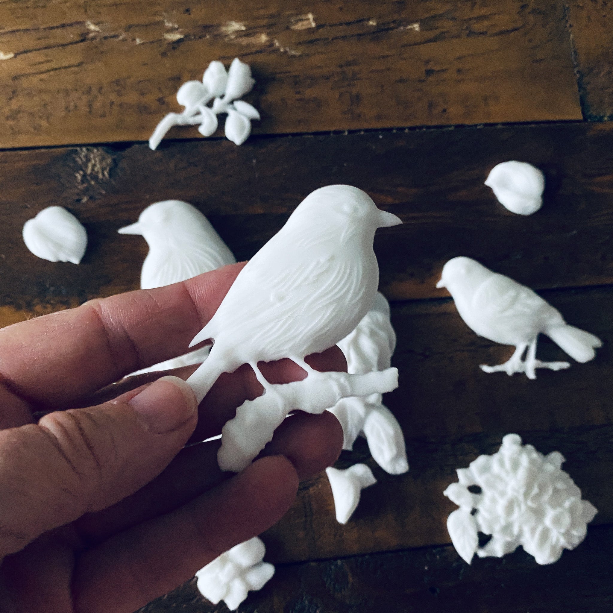A hand is holding a white resin casting against a wood background made from LeBlanche's Spring Birds silicone mold.