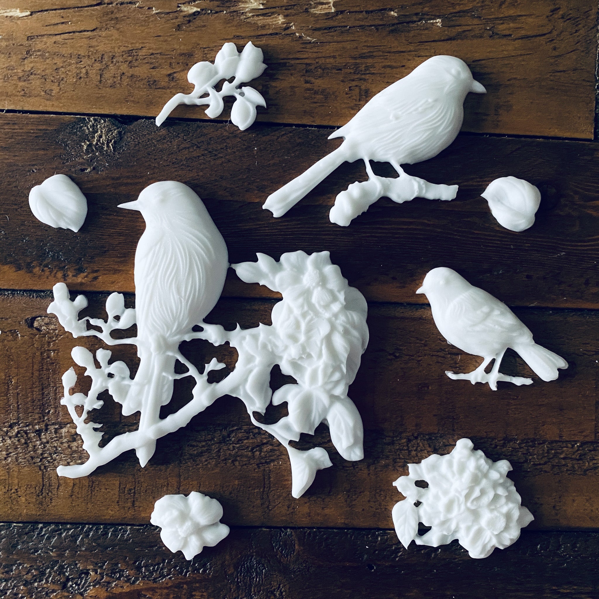 White resin castings against a wood background of 3 birds, 2 on branches, and some loose leaves and flowers made from LeBlanche's Spring Birds silicone mold.