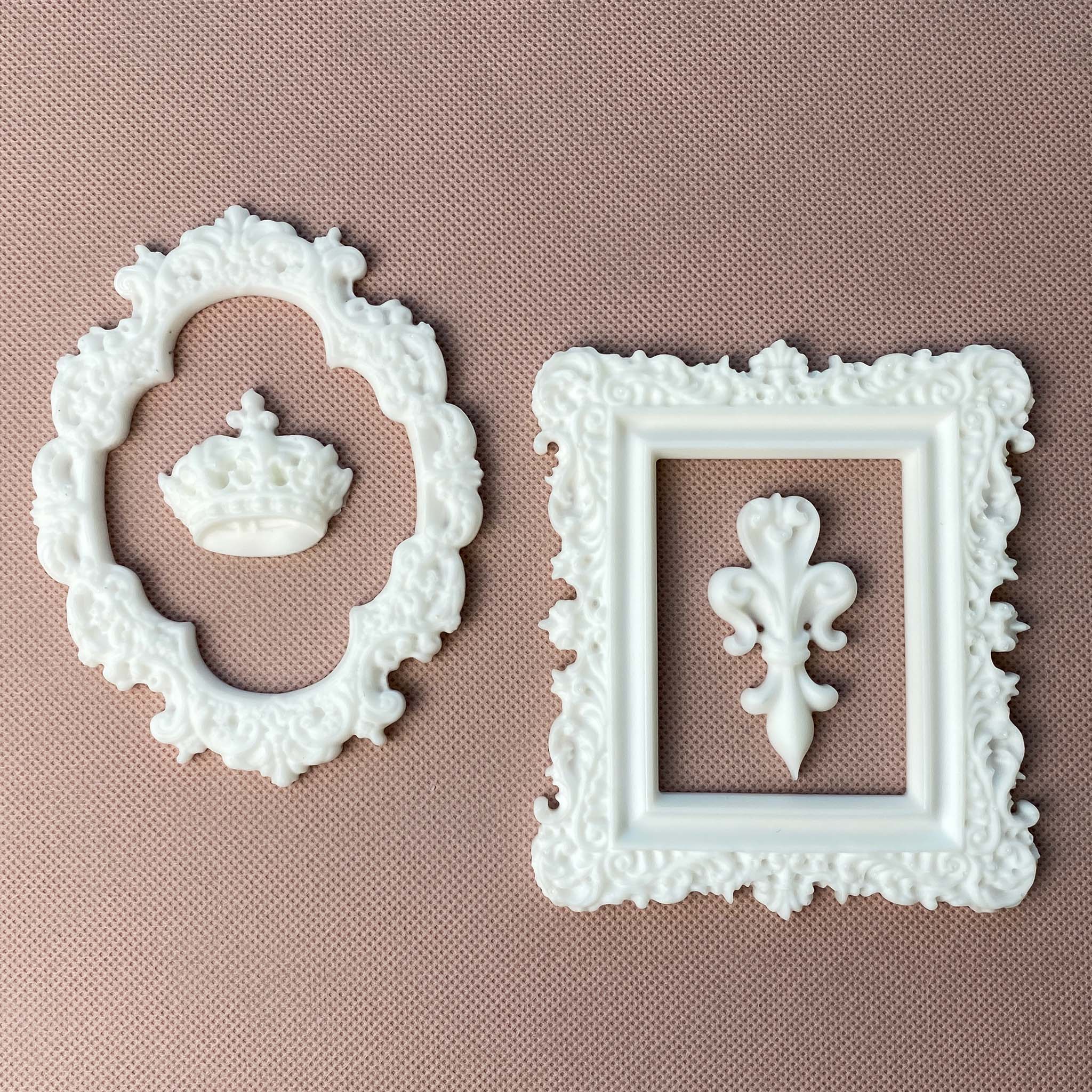 White resin castings of an ornate oval and rectangle frame are against a light beige material background. Inside the oval frame is a casting of a royal crown and inside the rectangle frame is a fleur de lis.