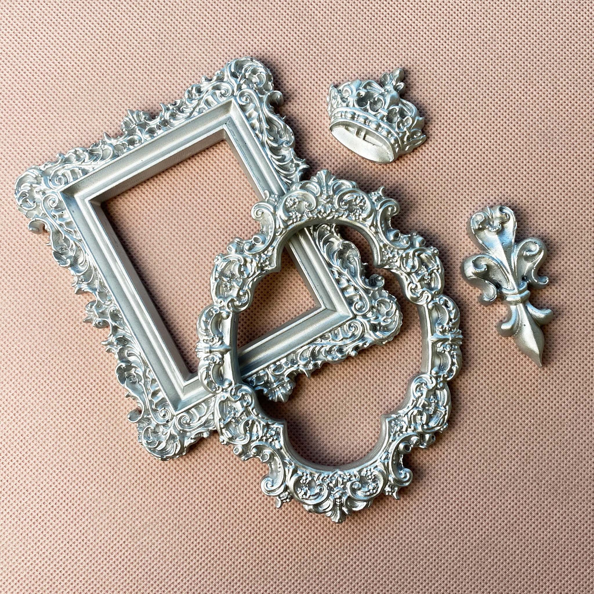 Silver colored castings of an ornate oval and rectangle frame, a small royal crown, and a fluer de lis are against a light beige material background.