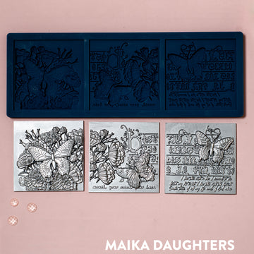 A blue silicone mold and silver colored castings of 3 tiles featuring butterflies, flowers and calligraphy are against a light pink background.