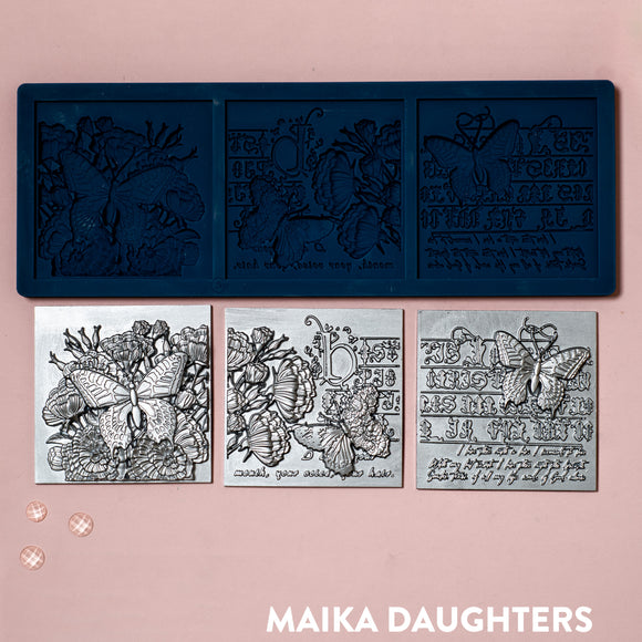A blue silicone mold and silver colored castings of 3 tiles featuring butterflies, flowers and calligraphy are against a light pink background.