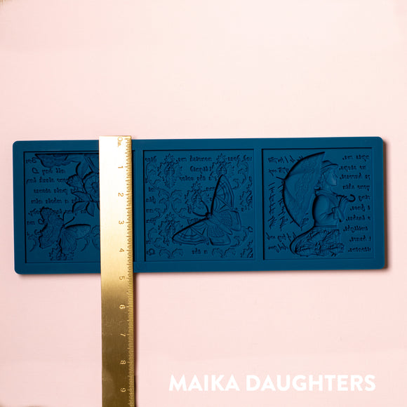 A blue silicone mold of 3 tiles featuring a woman holding an umbrella, butterflies, flowers, and script writing is against a light pink background. A gold ruler reading 4.8" height sits on the mold.