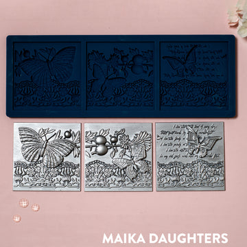 A blue silicone mold and silver colored castings of 3 tiles featuring butterflies, a lace border, and script writing are against a light pink background.
