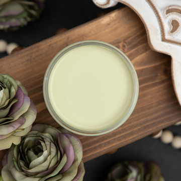 An arial view of an open container of Dixie Belle Paint Company’s Farmhouse Green Chalk Mineral Paint is on a wood plank surrounded by green with touches of purple silk roses.