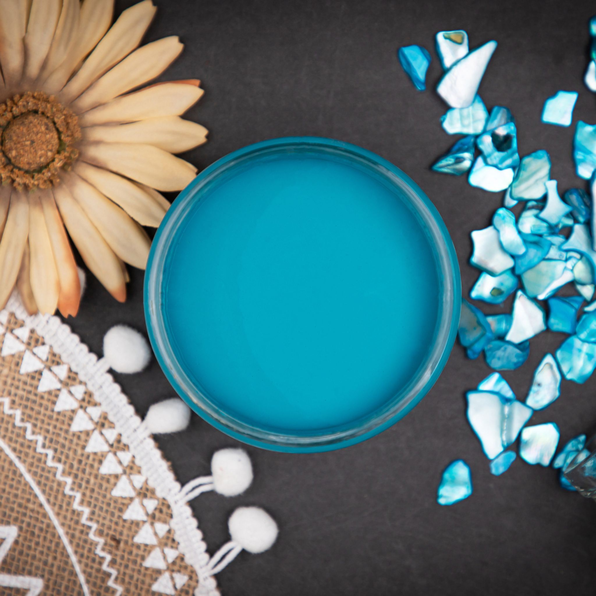 An arial view of an open container of Dixie Belle Paint Company’s Peacock Chalk Mineral Paint is surrounded by blue abalone shells, a daisy flower, and a burlap mat.
