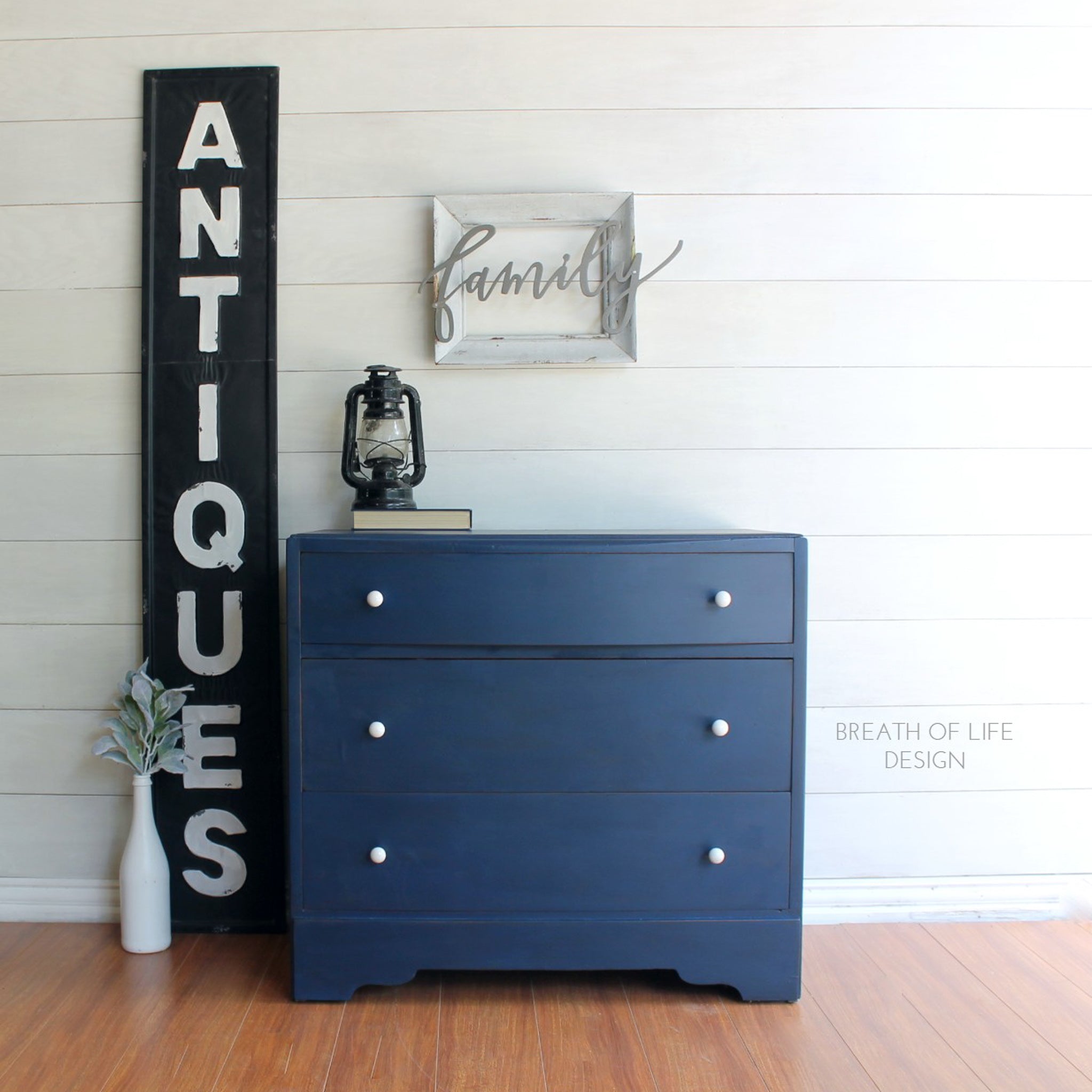 A 3-drawer dresser refurbished by Breath of Life Design is painted with Dixie Belle's Bunker Hill Blue chalk mineral paint. A large black and white sign that says "Antiques" sits to the left of the dresser.