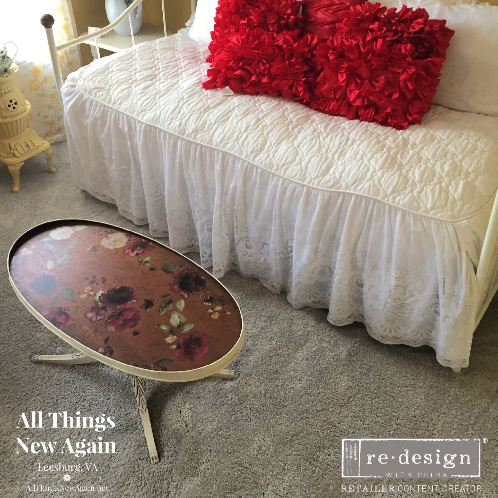 An oval vintage coffee table refurbished by All Things New Again is painted a light cream color on the legs and around the table top. The table top is a natural dark wood and features ReDesign with Prima's Midnight Floral transfer on it. A daybed with a white duvet cover and red pillow is also in the photo.