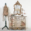 A vintage dresser is painted a light metallic gold and features the Chatellerault transfer on its drawers. A dressform is to the left of the dresser and an ornate gold framed mirror is above it.