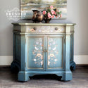 A vintage side table with storage refurbished by Brushed by Brandy is painted an ombre blend of light sage green down to a muted blue and features ReDesign with Prima's Albery transfer on its doors and drawer.