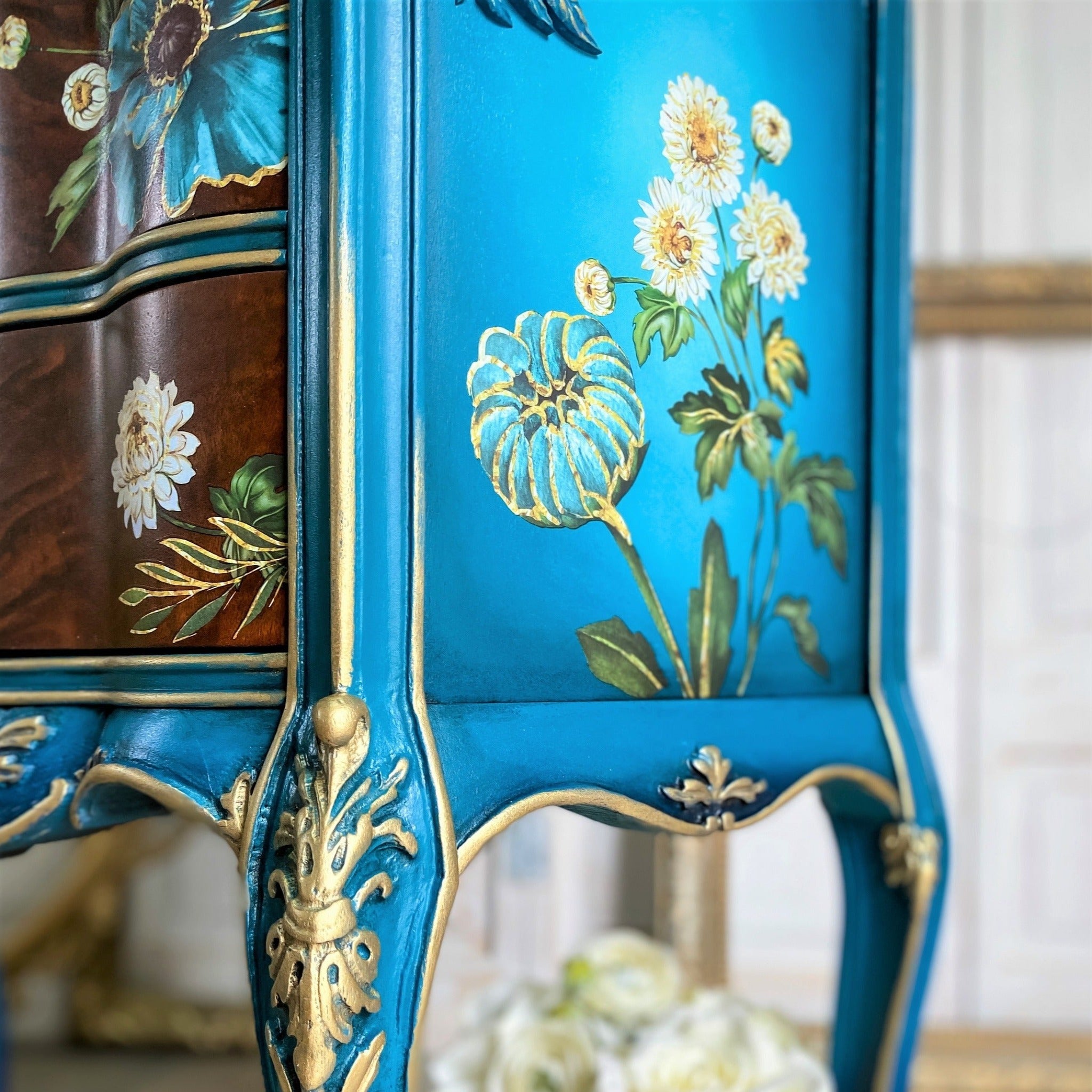 A close-up of a small 3-drawer nightstand refurbished by The Grandson's Brush is painted blue with gold accents and features Gilded Floral on the sides and on its natural wood stained drawers.