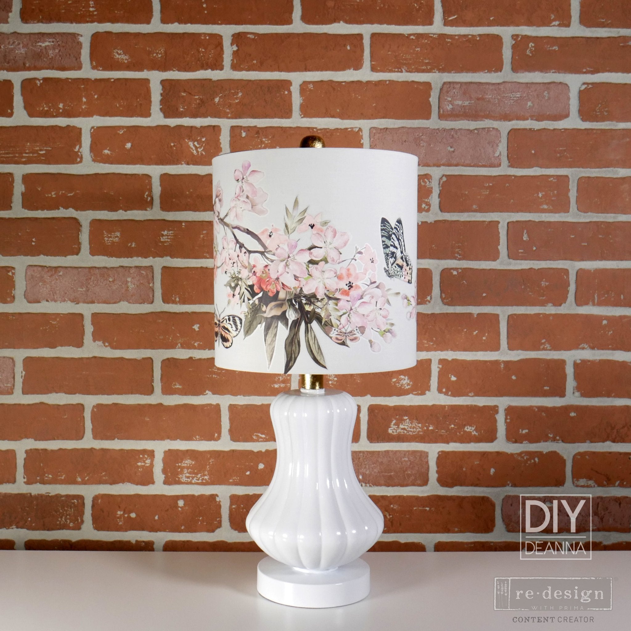 A white ceramic lamp refurbished by DIY Deanna is against a red brick wall backdrop and features ReDesign with Prima's Blossom Botanica on its white lamp shade.