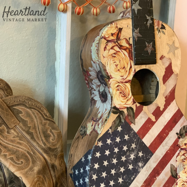 An acoustic guitar refurbished by Heartland Vintage Market features ReDesign with Prima's Sunflower Farms transfer on it along with an American Flag transfer.