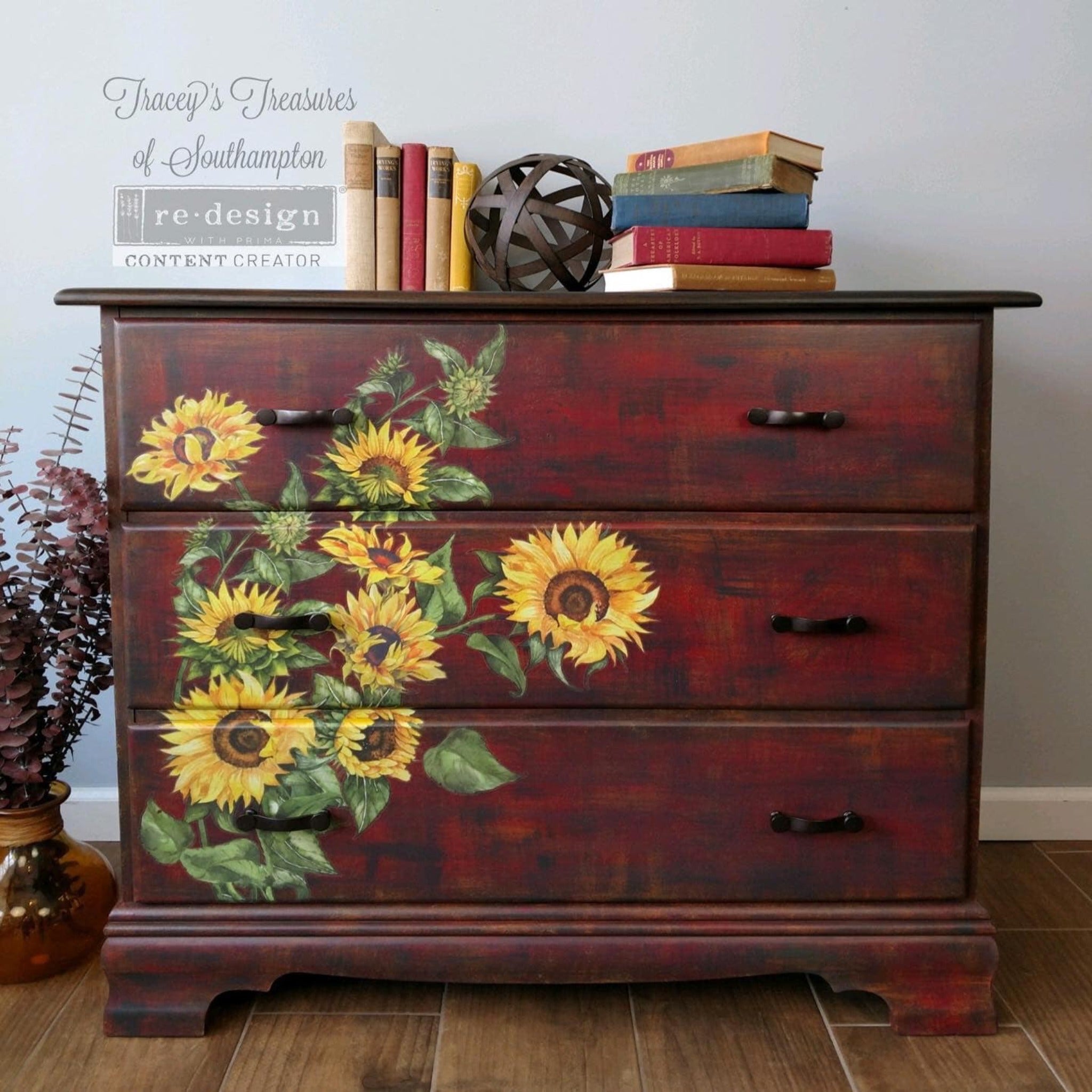A 3-drawer dresser refurbished by Tracey's Treasures of Southampton is painted a blend of red, black, and mustard yellow and features ReDesign with Prima's Sunflower transfer on the left side of the front of its drawers.