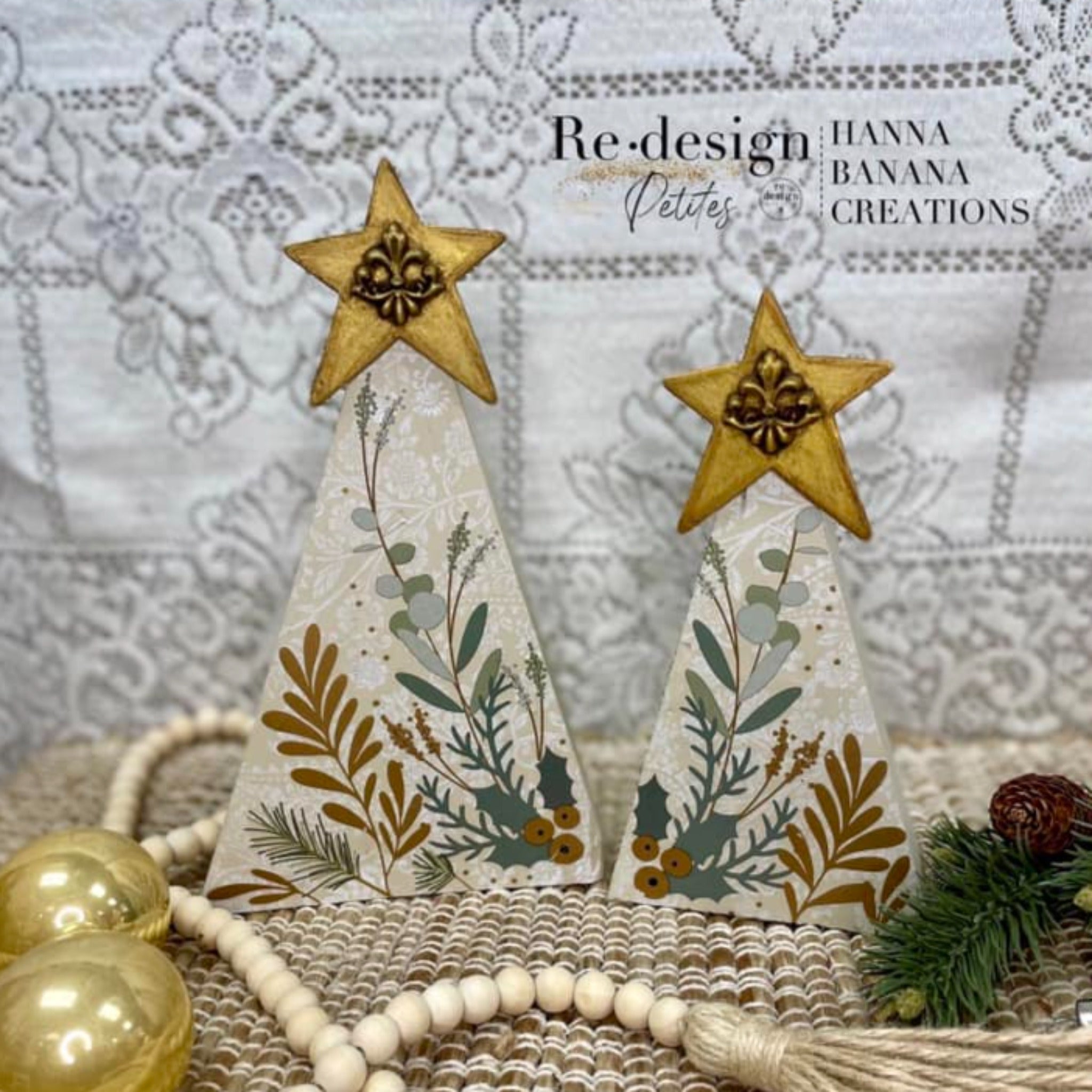 Two wood triangle Christmas tree crafts created by Hanna Banana Creations are painted beige with a white lace pattern background and feature ReDesign with Prima's Holiday Spirit small transfer on them. A large gold star with a decorative silicone mould casting are on the top of the trees.