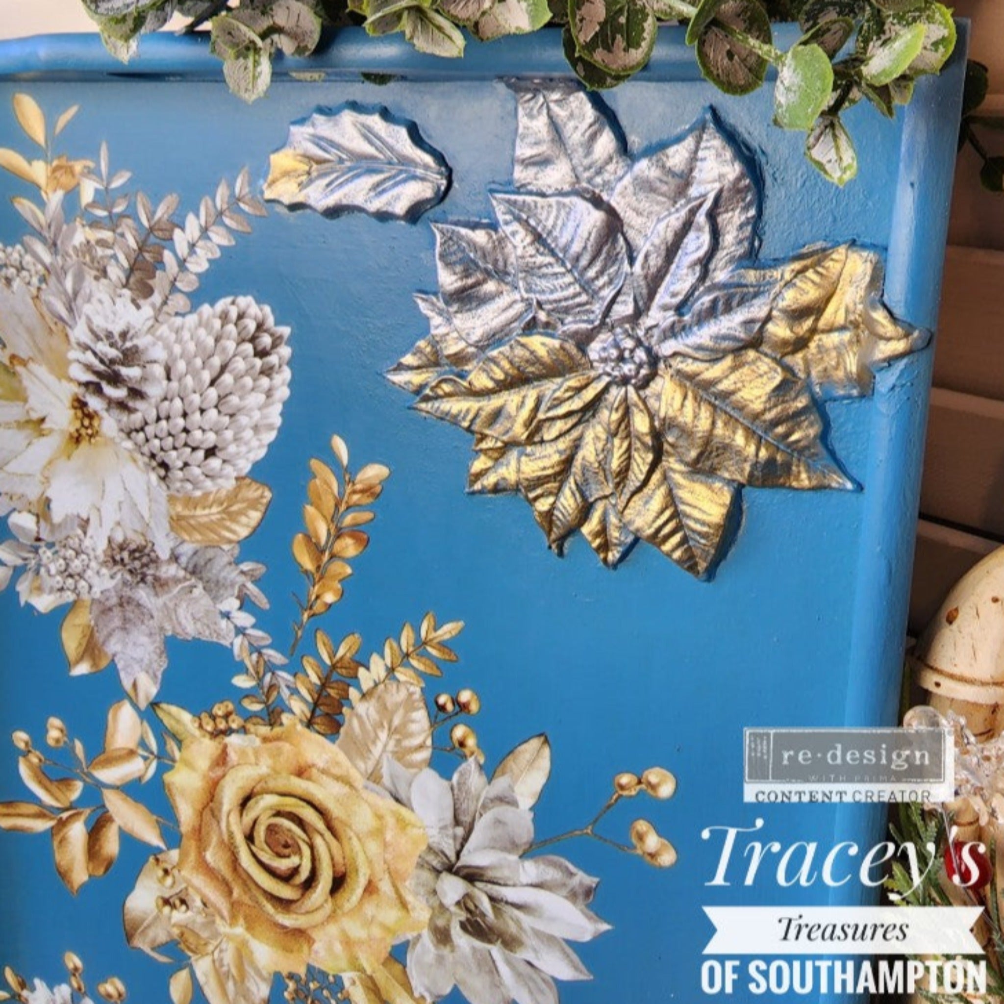 A close-up of a wood serving tray refurbished by Tracey's Treasures of Southampton is painted blue and features ReDesign with Prima's Perfect Poinsettia silicone mould casting in silver and gold colors at the top right inside the tray. ReDesign with Prima's A Gilded Moment floral transfer is also featured on the tray.