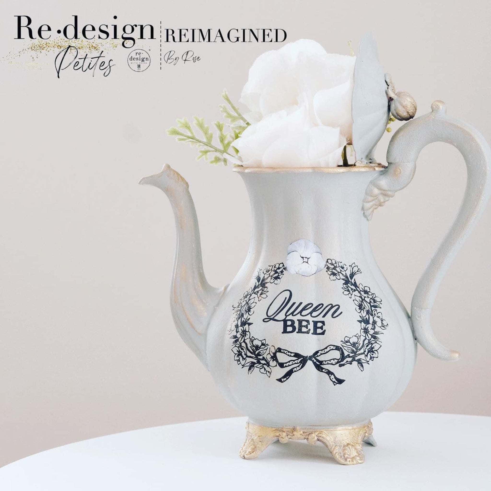 An antique white teapot with campagne gold accents refurbished by Reimagined by Rose features ReDesign with Prima's French Labels small transfer that says Queen Bee with a floral wreath around it.