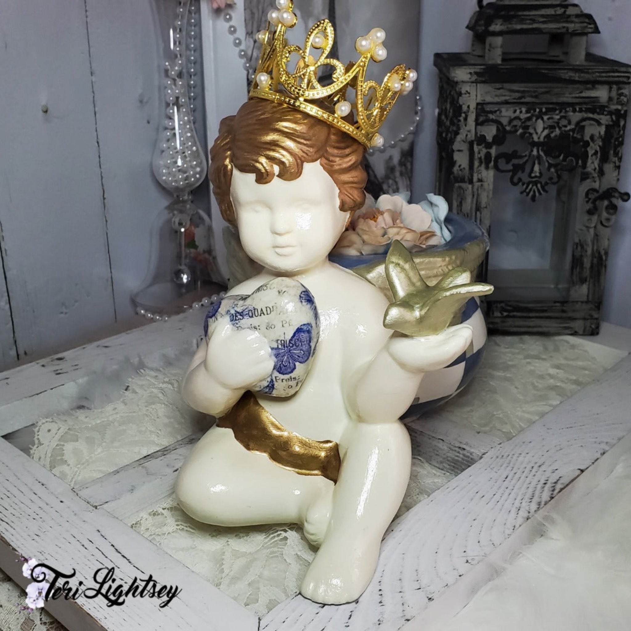 A small ceramic child statue is holding a heart that features Decoupage Queen's Blue Flight rice paper in one hand and a gold bird in the other hand.