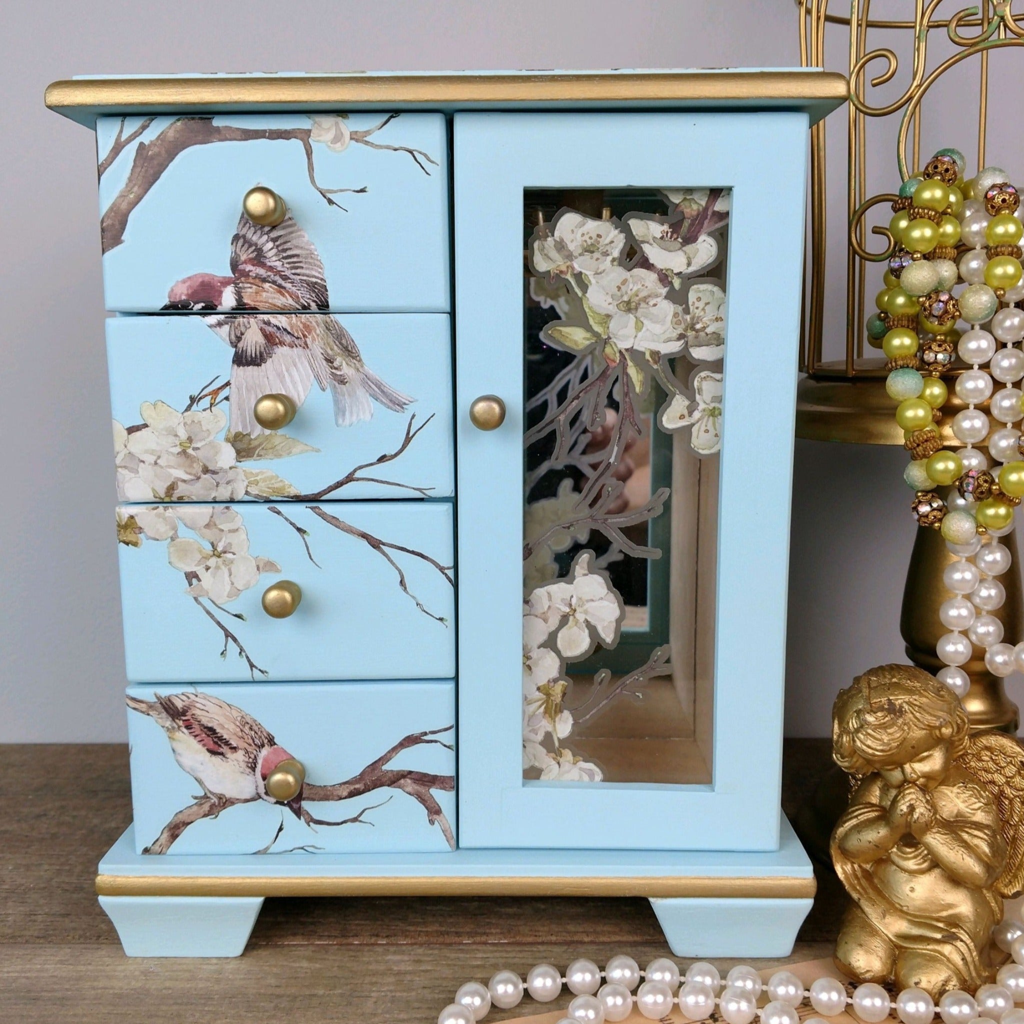 A small tabletop jewelry armoire is painted light blue with gold accents and features ReDesign with Prima's Blossom Flight transfer on it.