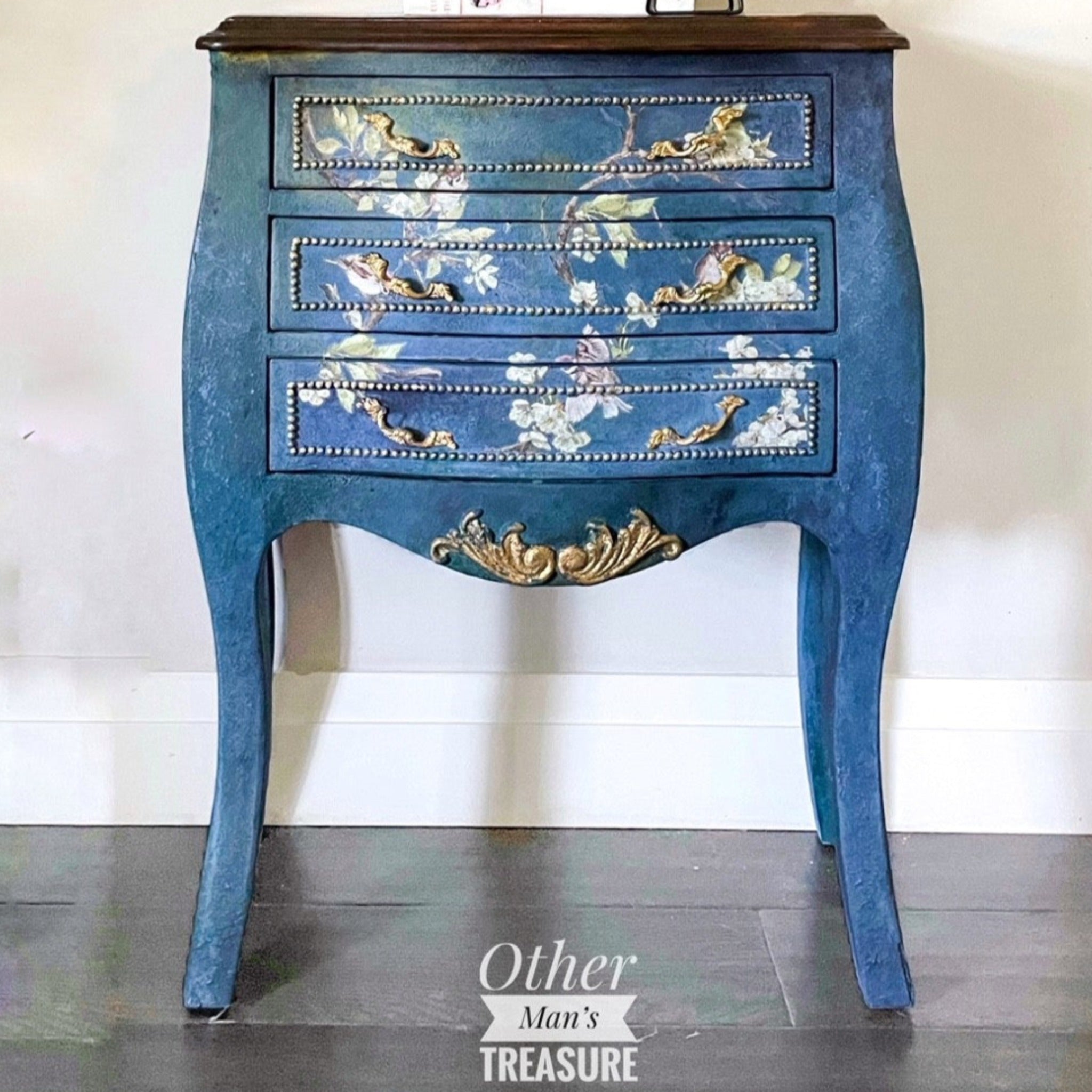 A vintage Bombay style nightstand refurbished by Other Man's Treasure is painted blue with gold accents and features ReDesign with Prima's Blossom Flight transfer on its small 3 front drawers.