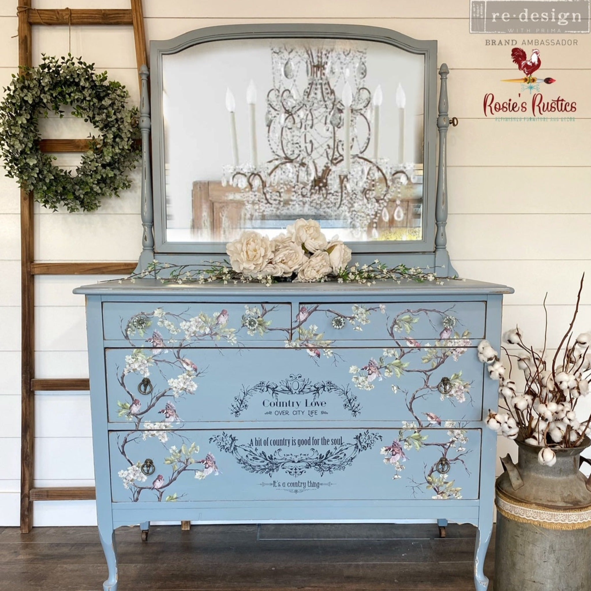 A vintage dresser with attached mirror refurbished by Rosie's Rustics is painted light blue and features ReDesign with Prima's Blossom Flight transfer on its drawers.