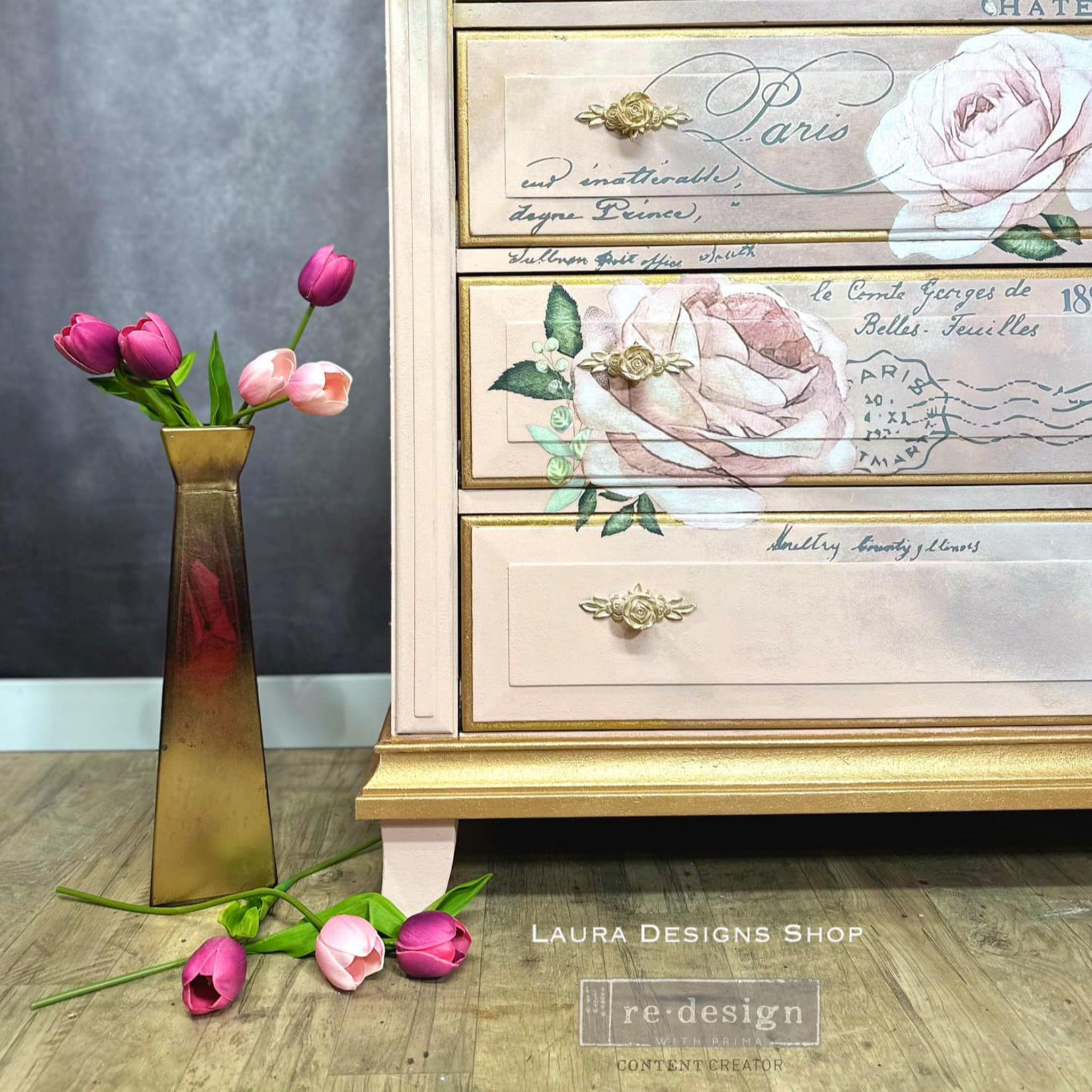 A close-up of a vintage dresser refurbished by Laura Designs Shop, a ReDesign with Prima Content Creator, is painted pale pink and gold accents and features the Chatellerault transfer on its drawers.