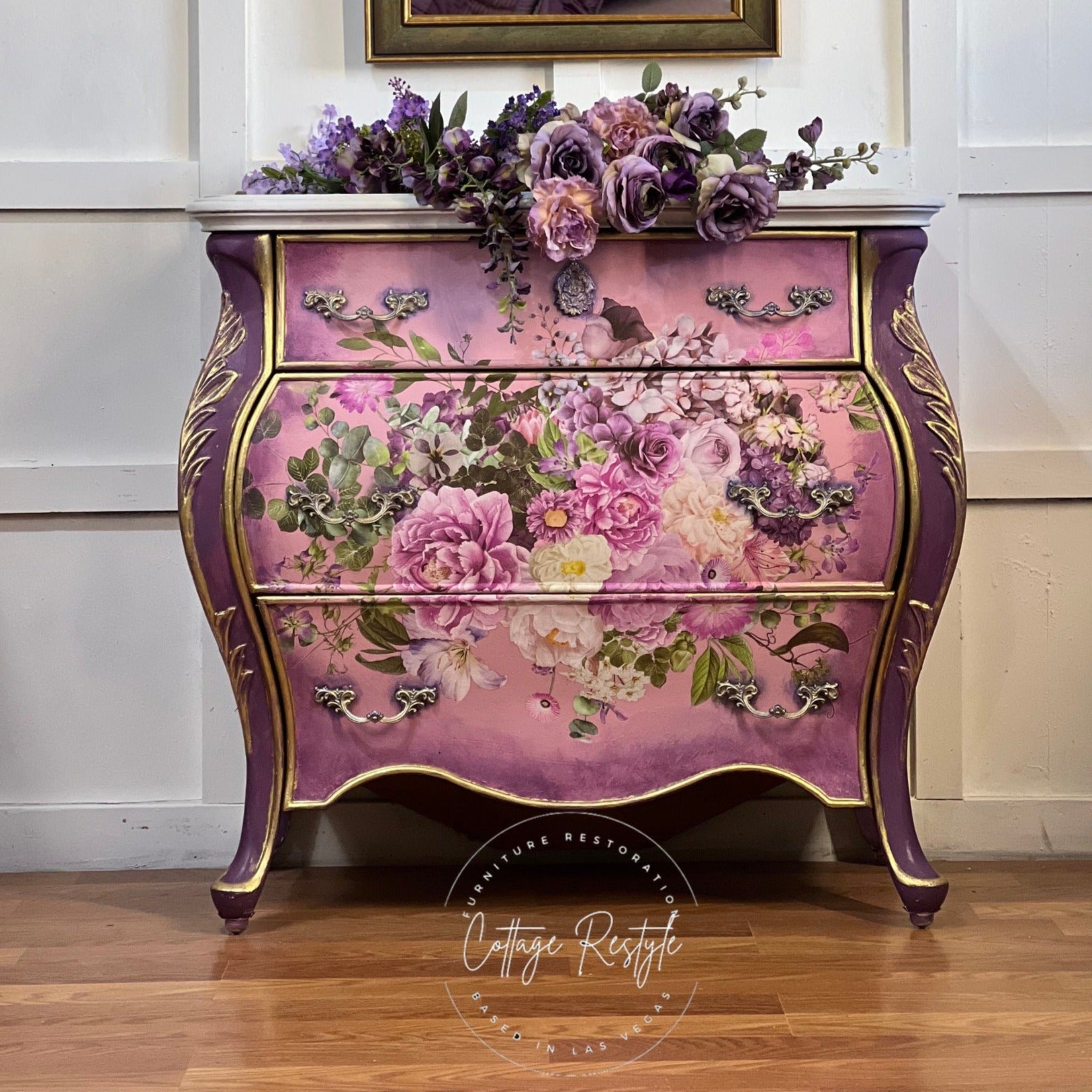 A vintage Bombay 3 drawer dresser refurbished by Cottage Restyle is painted purple with gold accents on the legs that run up the sides of the dresser and a blend of light pink and purple on the drawers. The center of the drawers feature ReDesign with Prima's Kacha Morning Purple transfer on them.