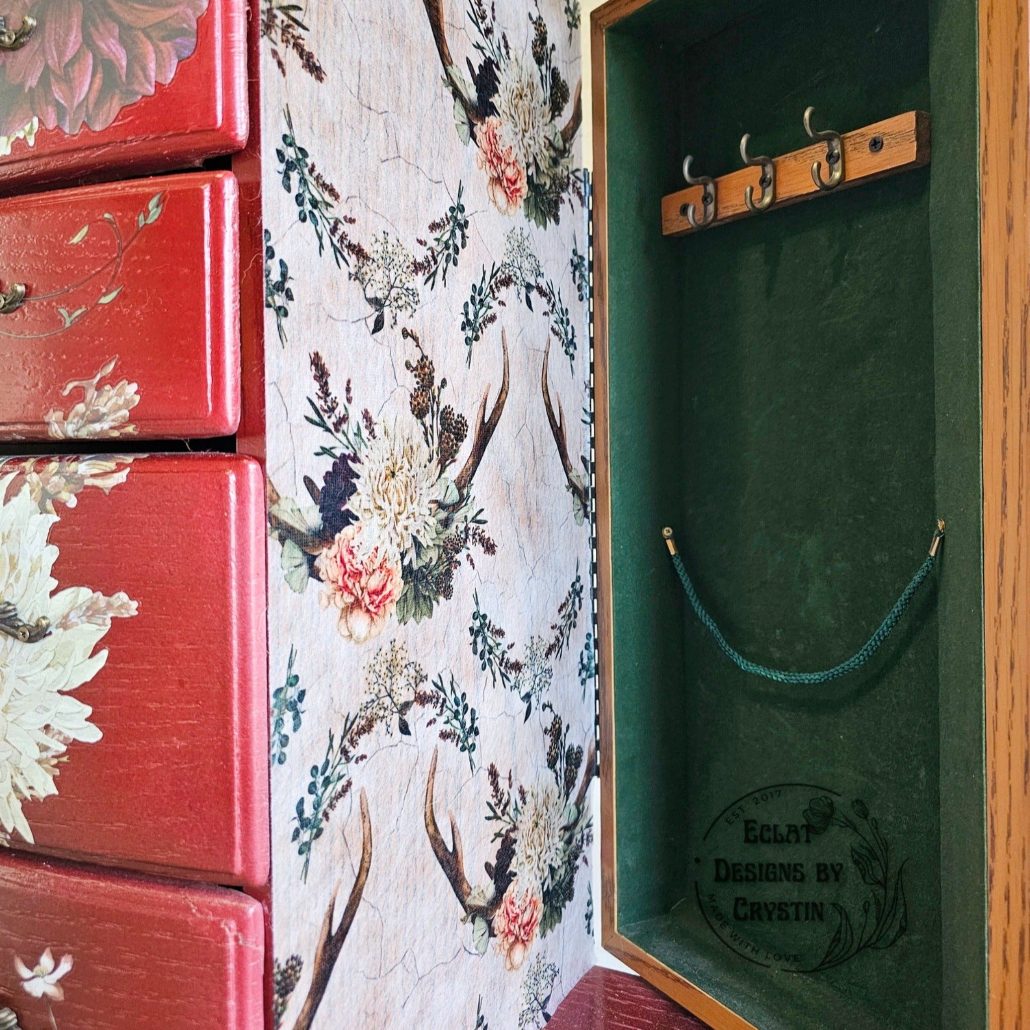 A close-up corner angle view of a vintage jewelry armoire refurbished by Eclat Designs by Crystin is painted red. The side door is open and shows green velvet inside the door and features ReDesign with Prima's Cedar Creek tissue paper on the side of the armoire.