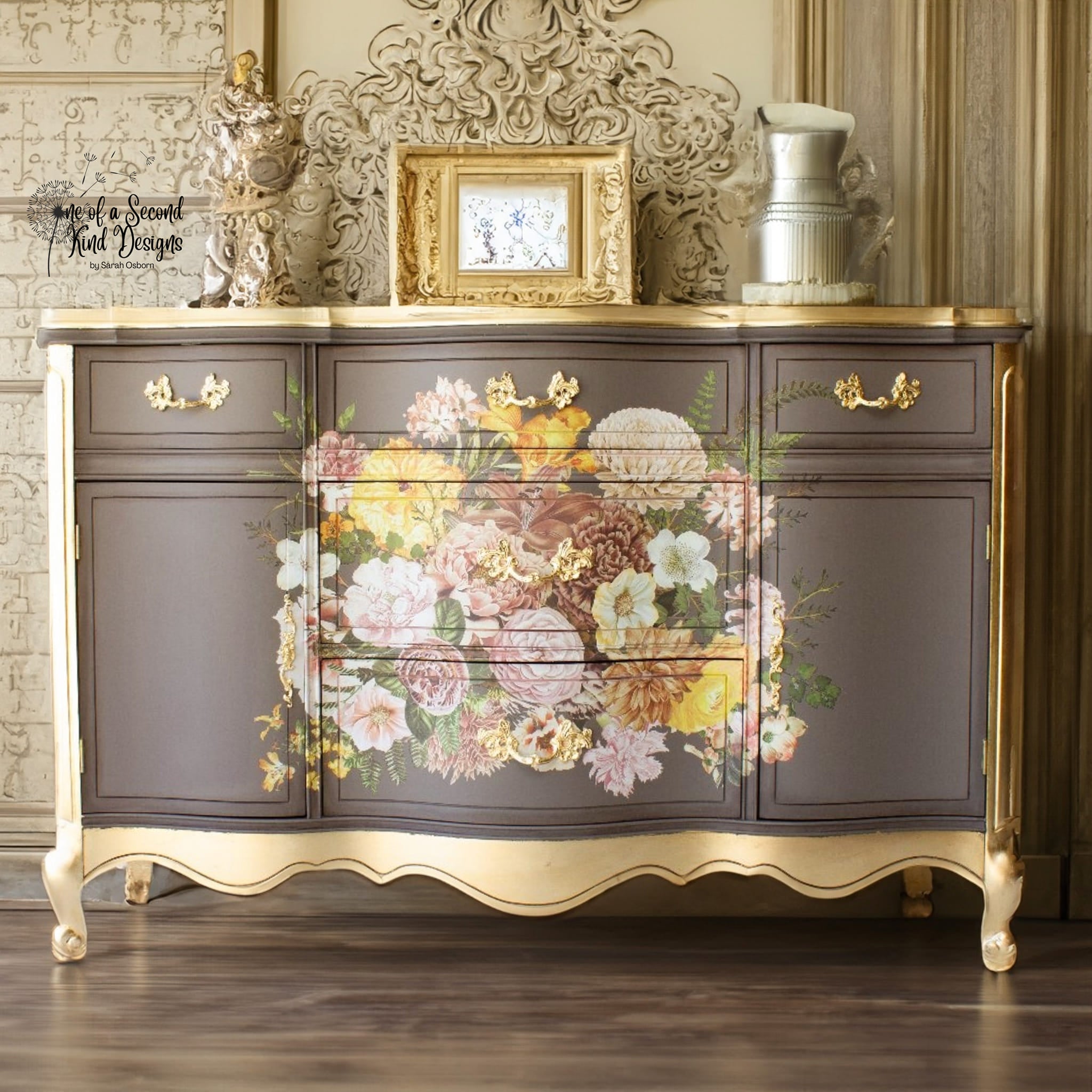 A vintage dresser with 5 drawers and 2 doors refurbished by One of a Second Kind Designs is painted a soft light brown with gold accents and features ReDesign with Prima's Kacha Woodland Floral transfer on the center of the center drawers.