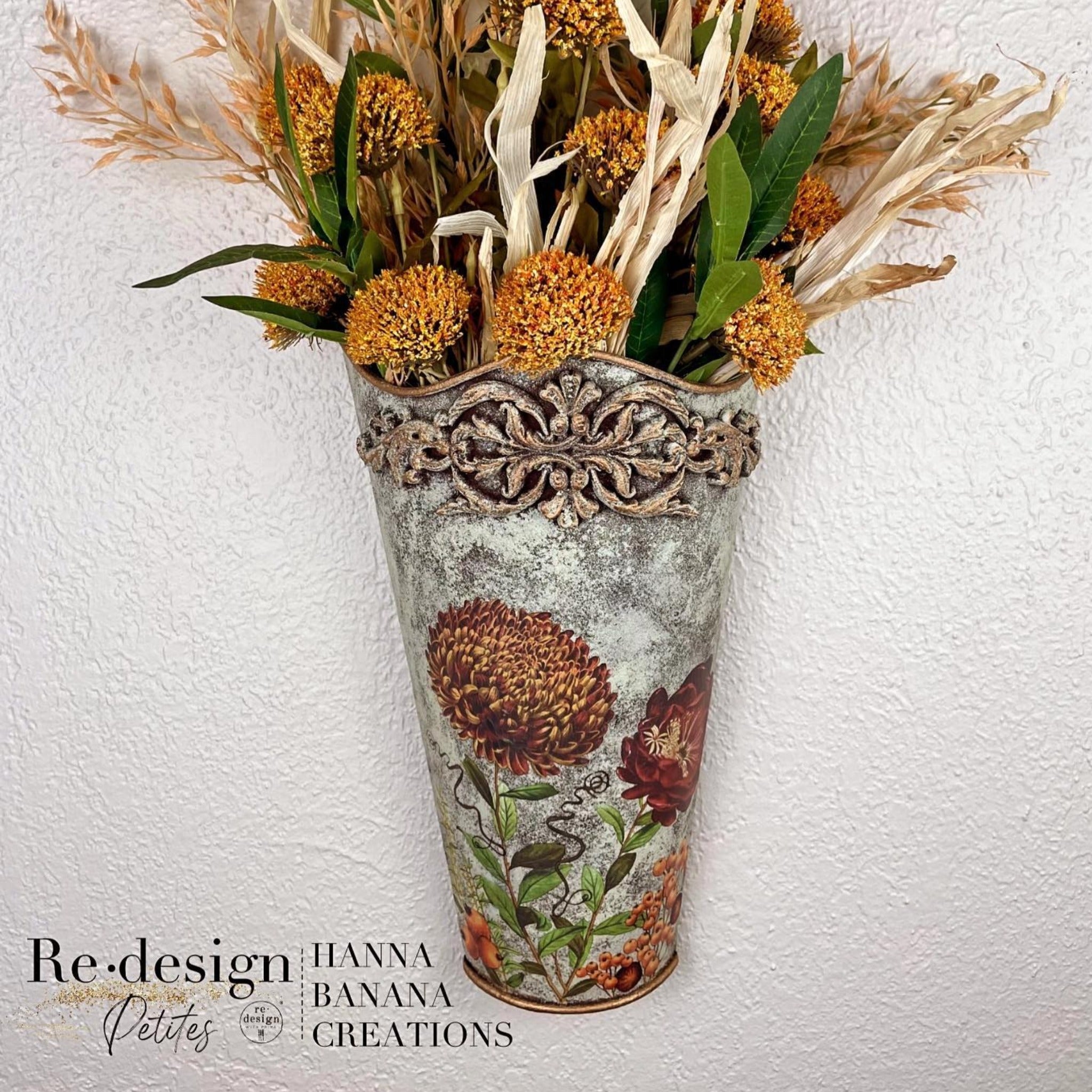 A metal can vase refurbished by Hanna Banana Creations is painted a concrete-like blend is greys and features ReDesign with Prima's Seasonal Splendor small transfer on it. A bouquet of dried Autumn flowers are in the vase.